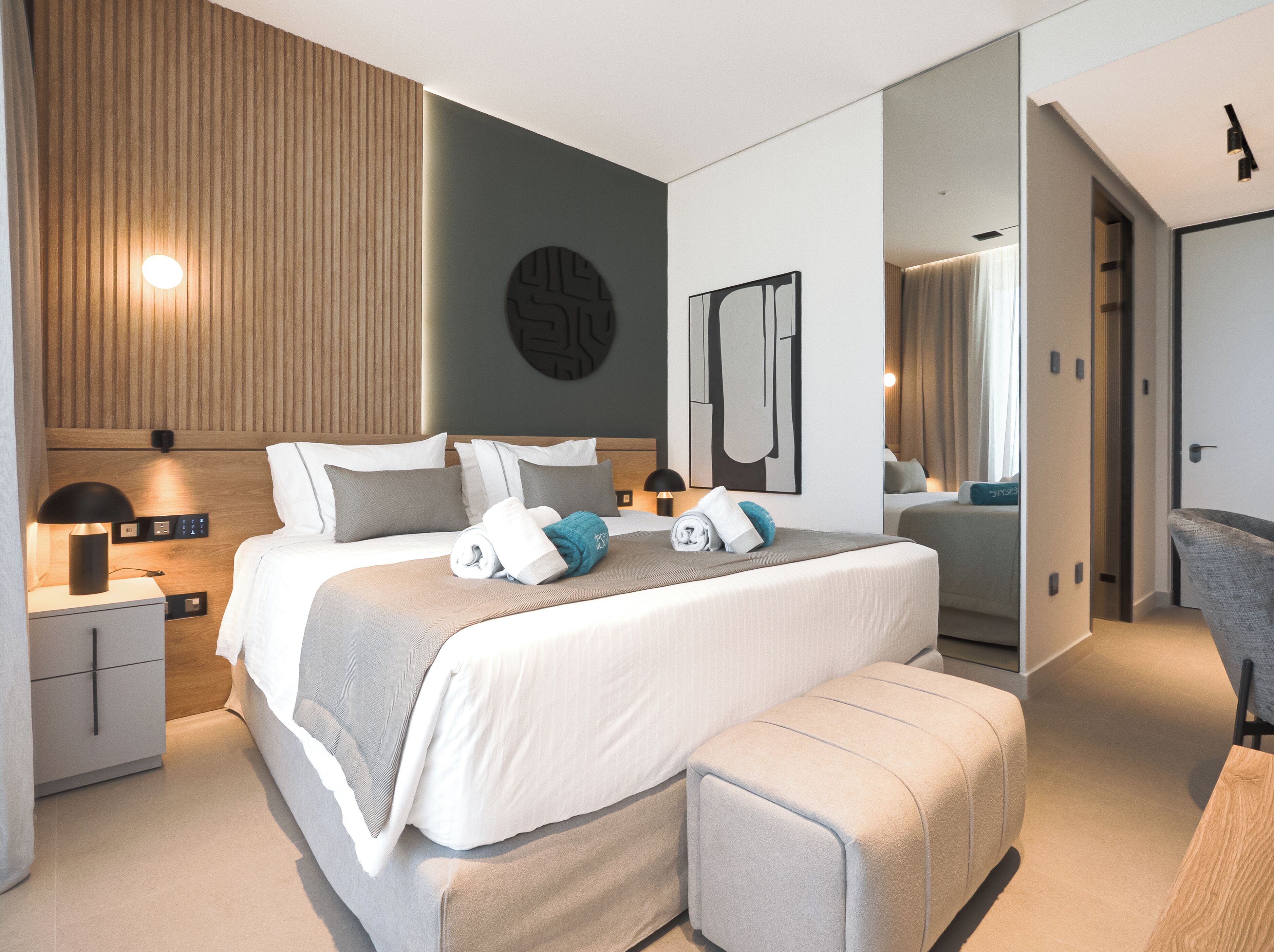 A modern hotel room with a large bed, bedside tables, a mirror, and a view of the adjacent room.