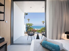 Habitación de hotel moderna con balcón y vista al mar, una cama con toalla azul, y palmeras y edificios a la vista.