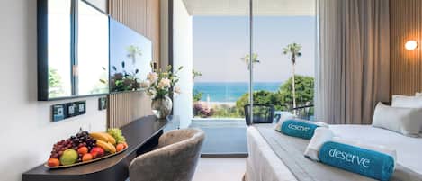 A hotel room with a bed, a desk with fruit, a chair, a large window with a view of the ocean, and a mirror on the wall.