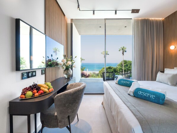 Habitación de hotel con cama, escritorio con fruta, una silla, ventana grande con vista al océano y espejo en la pared.