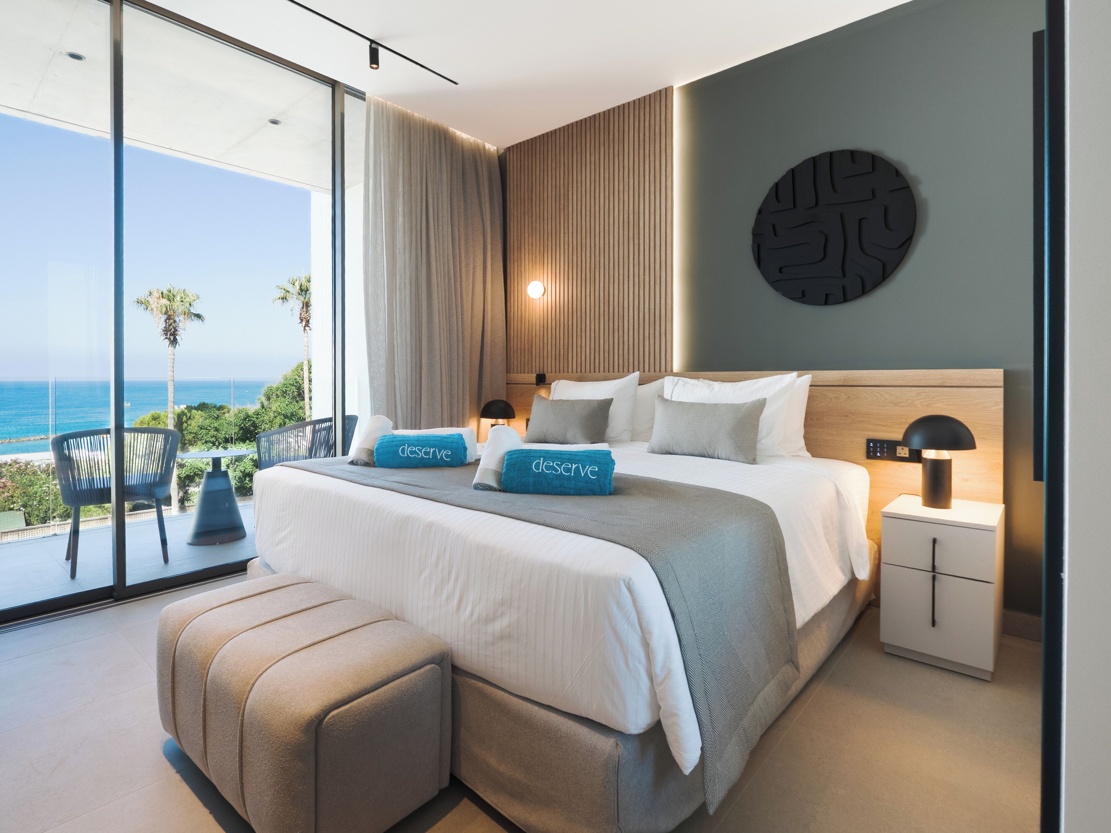 A modern bedroom with a large bed, a nightstand, a wall-mounted lamp, and a view of the ocean.
