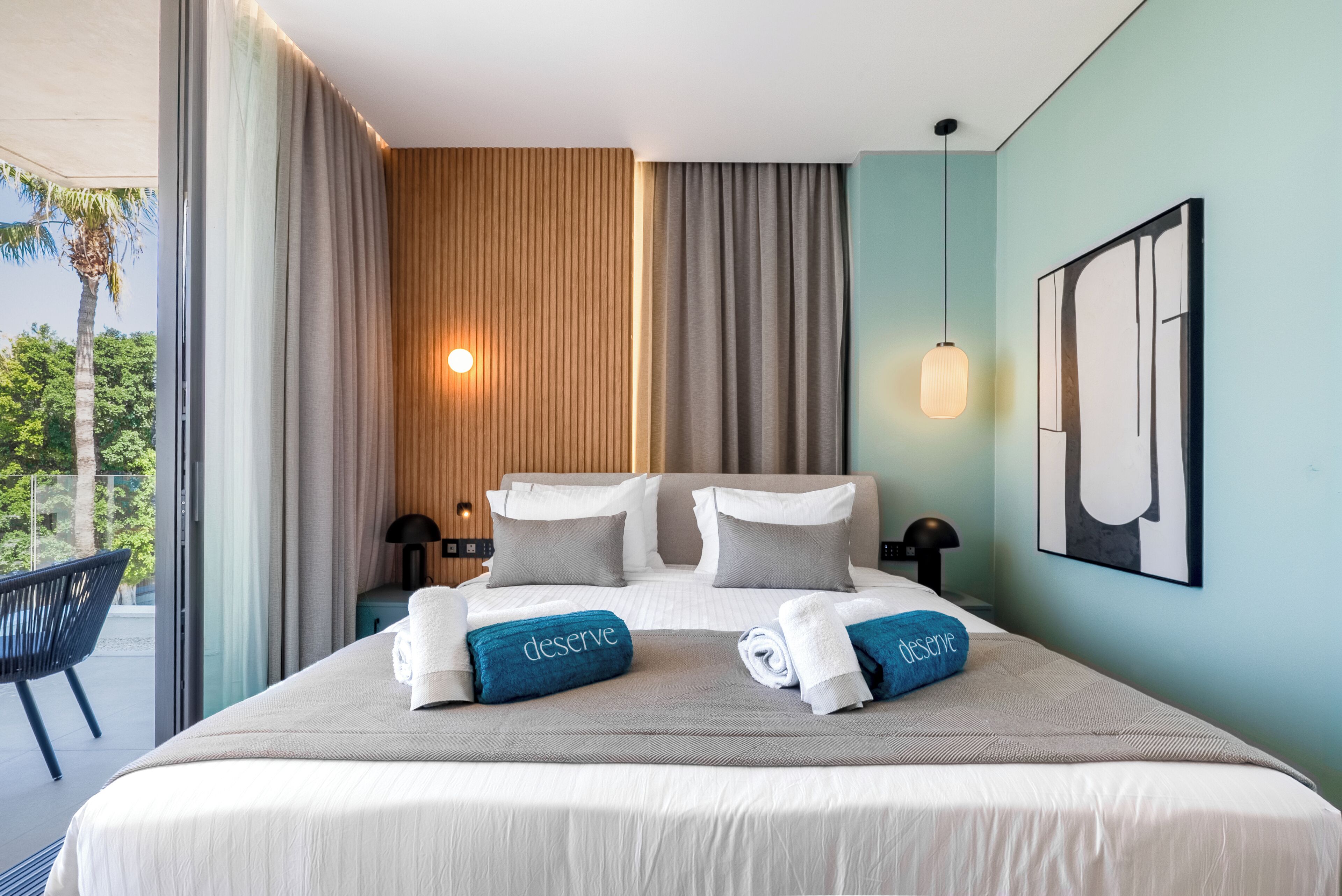 Un cuarto de hotel con una cama grande, dos lámparas de noche, vista a palmeras y un cuadro en la pared.
