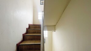 Un escalier étroit avec des marches en bois et une rampe métallique, menant à un palier doté d’une petite fenêtre.