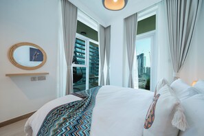 A modern hotel room with a large bed, a view of a cityscape through floor-to-ceiling windows, and a round mirror on the wall.