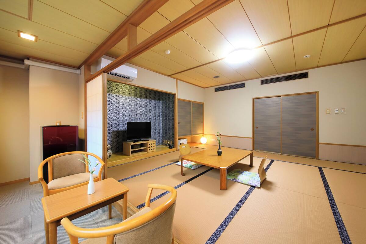 Une pièce de style japonais traditionnel, avec un sol recouvert de tatamis, une petite table en bois, deux chaises, un téléviseur et des portes coulissantes.