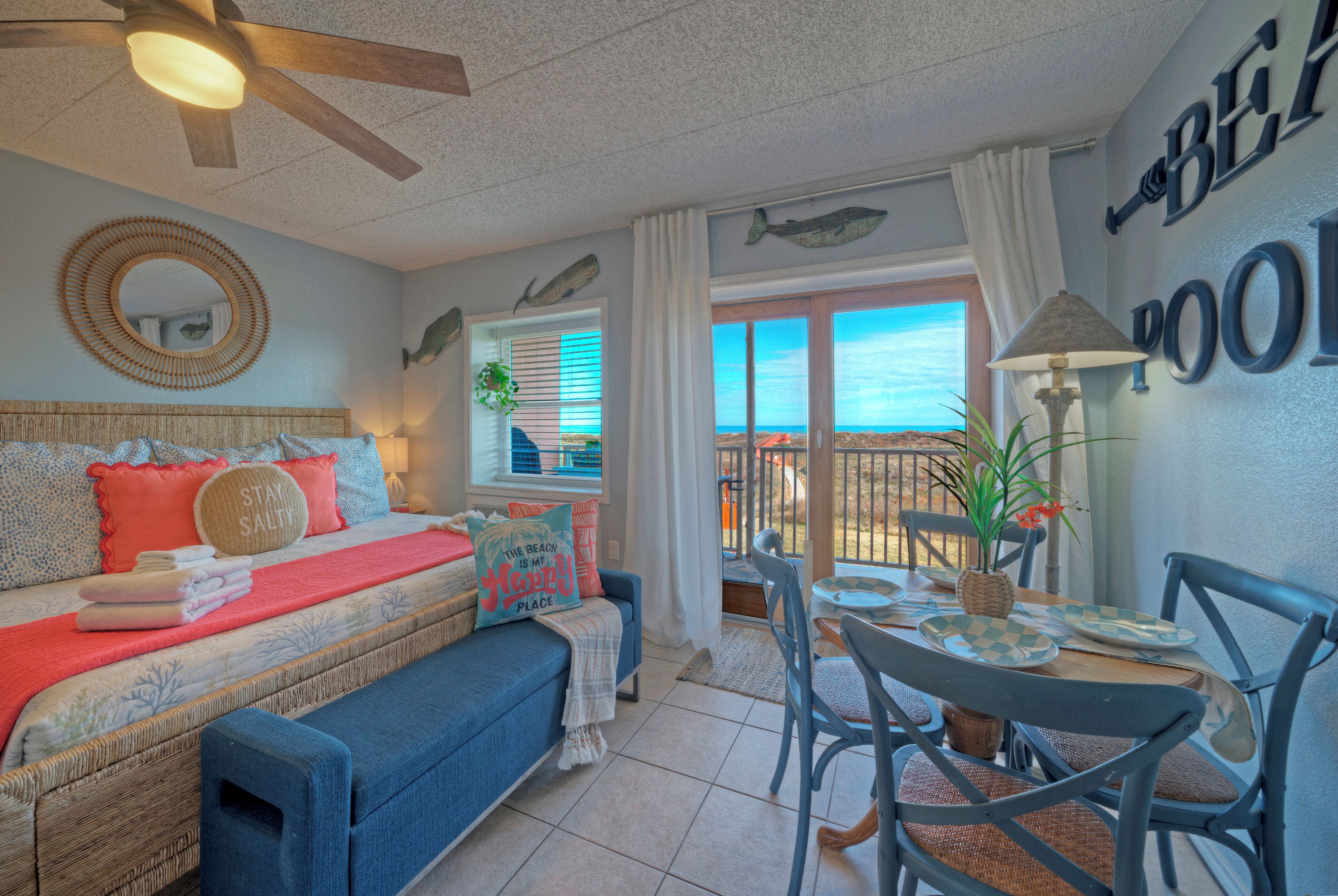 A bedroom with a bed, a window with a view, a ceiling fan, and a dining area with chairs and a table.