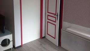 Een kamer met een witte deur met rode accenten, een wit kastje met een zwart voorwerp erop, en een witte wasbak met een geperforeerd paneel erboven.