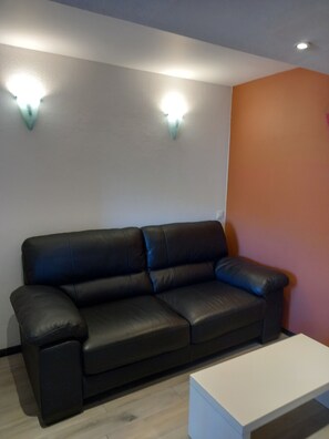 A dark leather sofa against a white wall with two wall-mounted lights, a white side table, and a wooden floor.