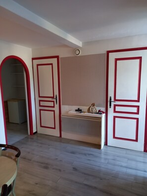 A room with a kitchenette, a door with a red frame, a white ceiling, and a wooden floor.