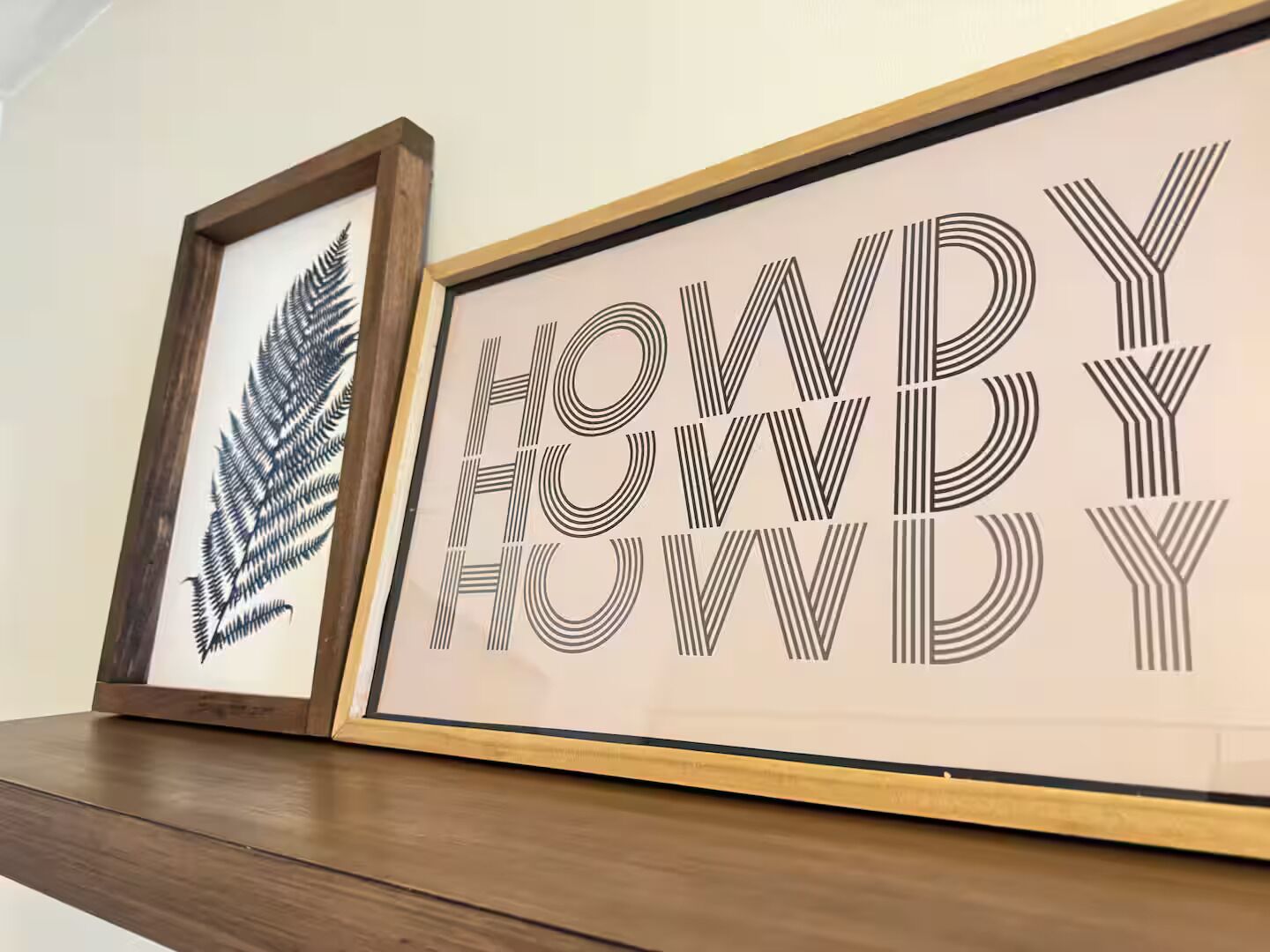 Two framed artworks on a wooden shelf. One artwork features a black fern leaf, and the other displays the word "HOWDY" in a stylized font with lines.