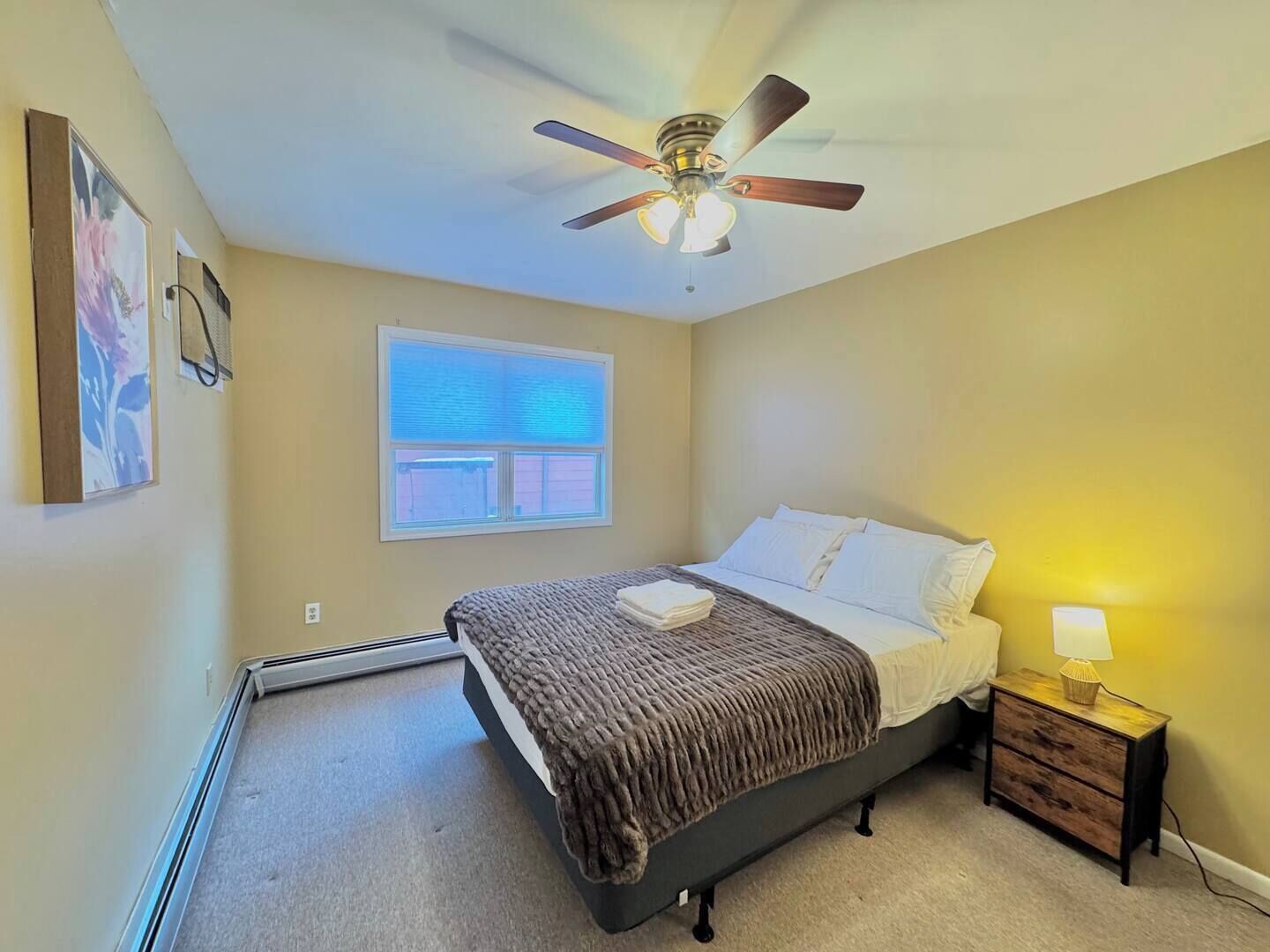 Un dormitorio con cama, ventilador de techo, ventana con persianas, mesita de noche con lámpara y aire acondicionado de pared.