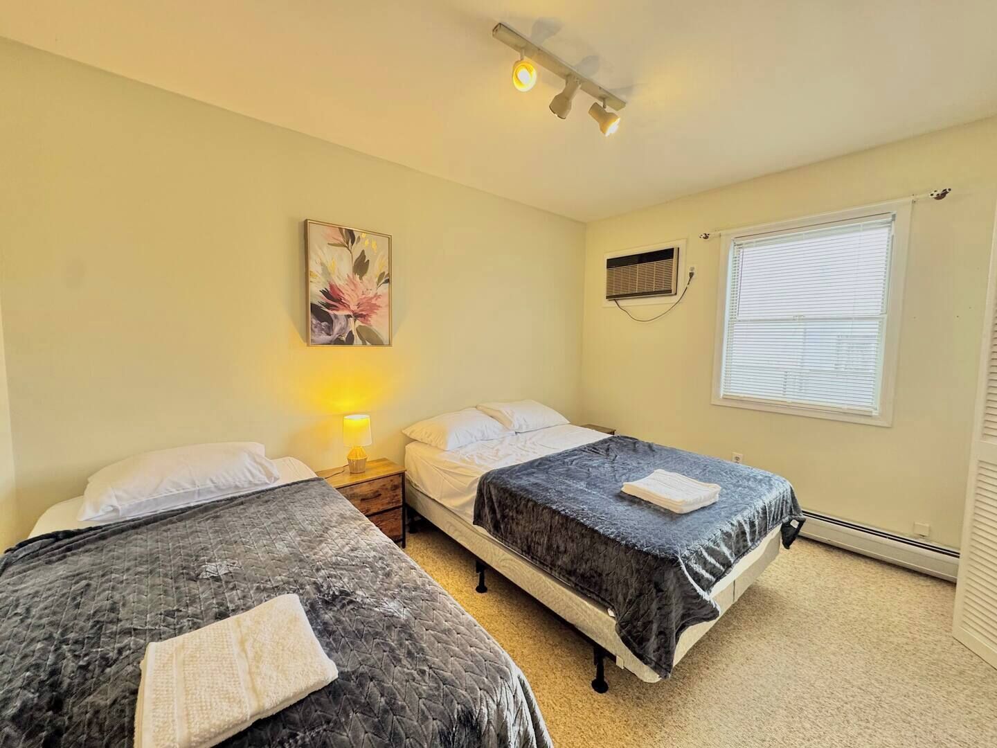 A bedroom with two beds, a nightstand with a lamp, a wall-mounted air conditioning unit, and a window with blinds.