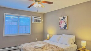 A bedroom with a bed, two bedside lamps, a ceiling fan, an air conditioning unit, and a window with blinds.