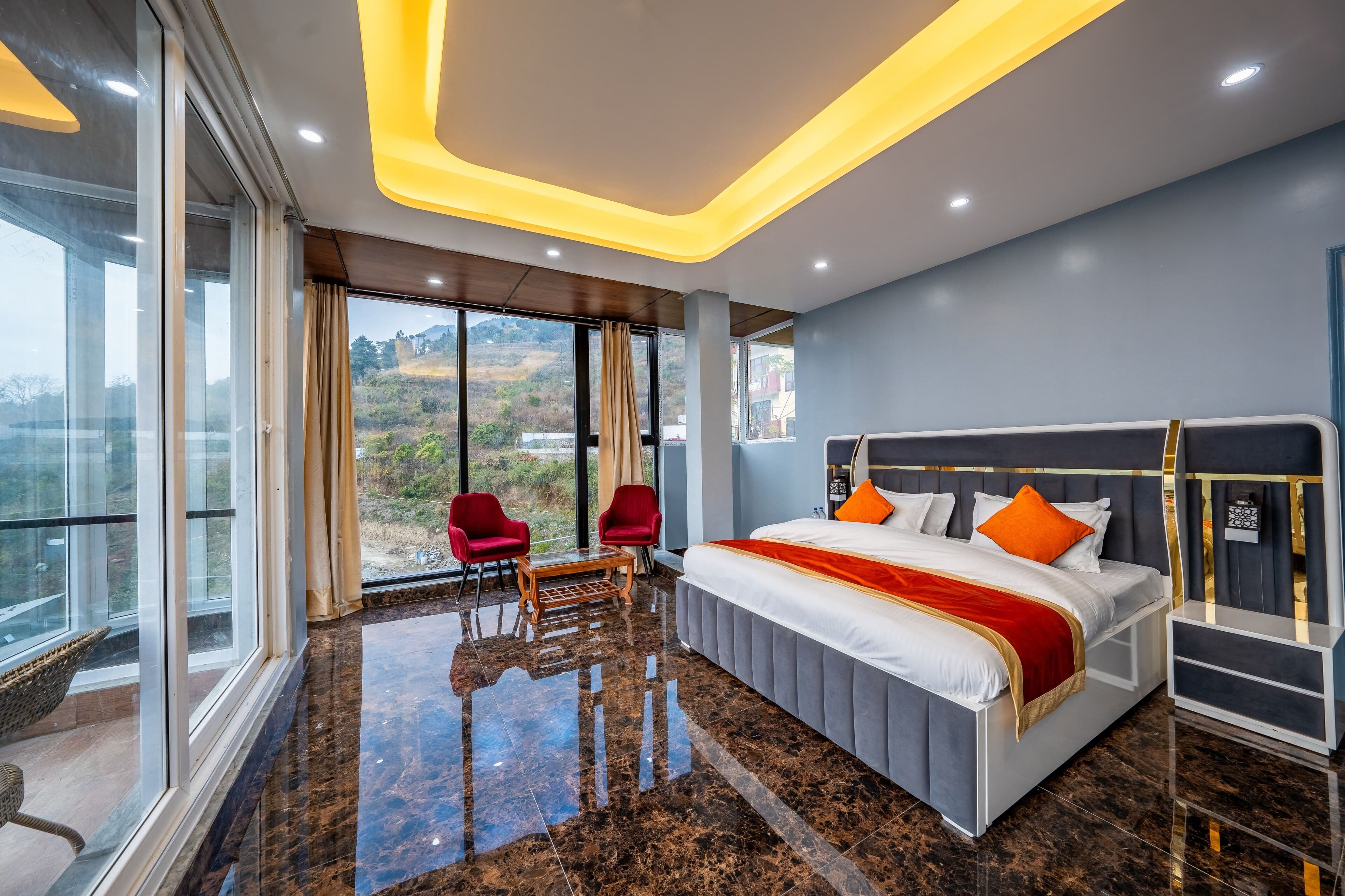 A modern hotel room with a large bed, a glass table, two red chairs, and a view of the outdoors.
