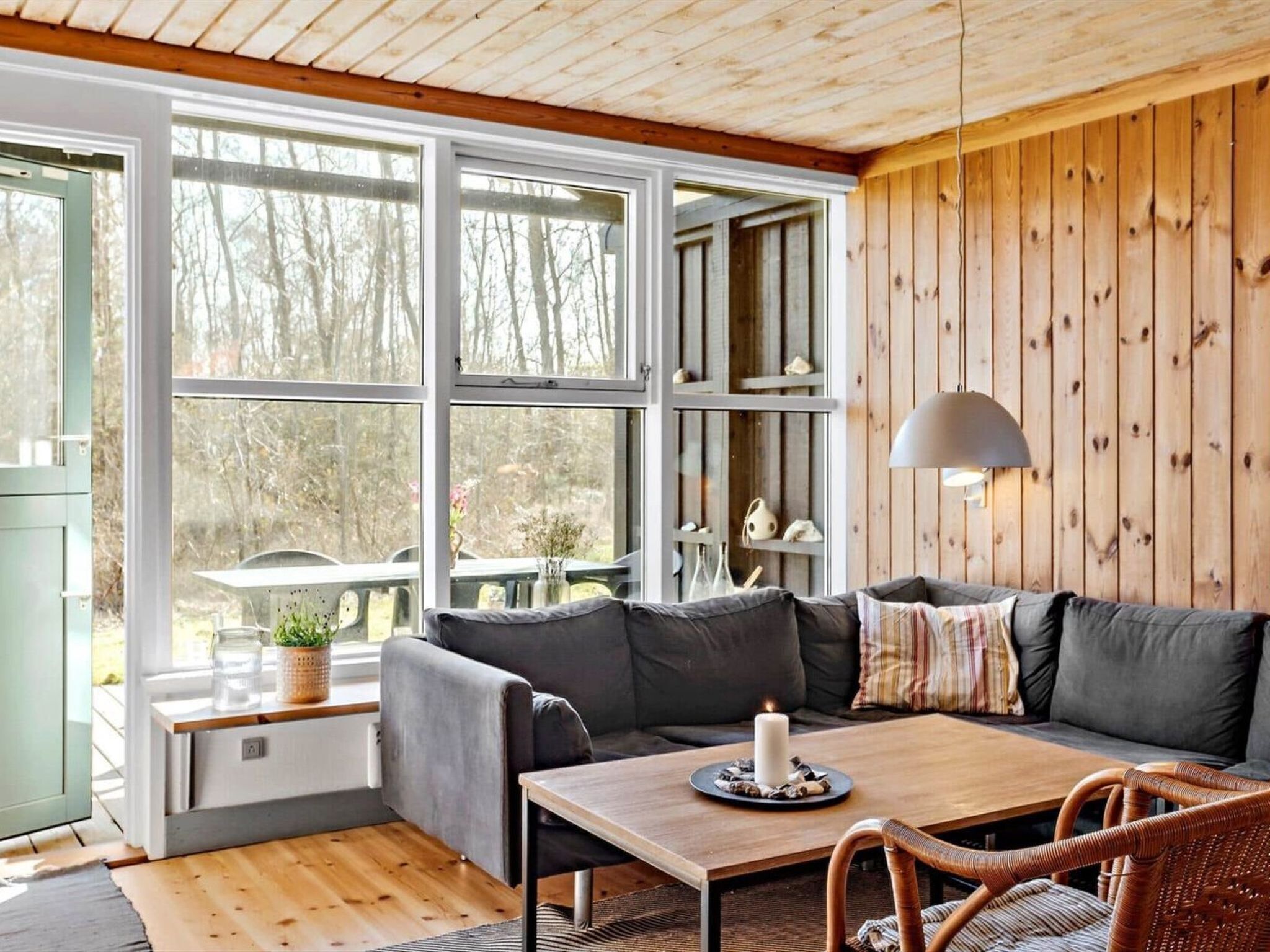 A cozy cabin interior with a wooden ceiling, large windows, a sofa, a table, and a wicker chair.