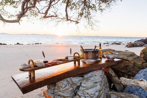 A wooden table set up on a beach with various food items and drinks, overlooking the ocean.