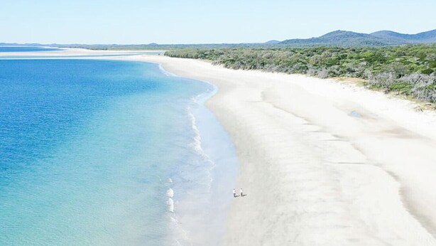 Una amplia playa de arena con agua azul cristalina y una cadena montañosa distante.