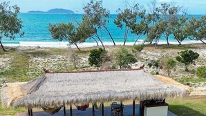 Una estructura de techo de paja con terraza de madera y zona de estar, con vista a una playa con árboles y el mar al fondo.
