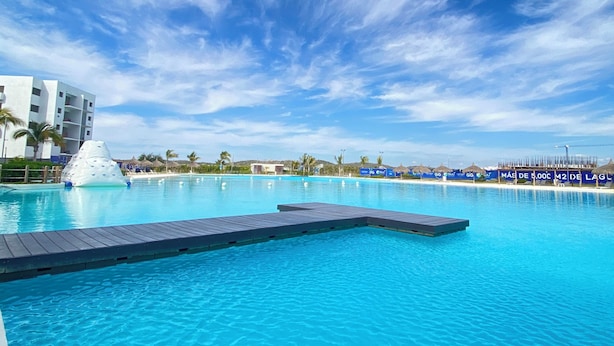 A clear blue sky with clouds, a swimming pool with a wooden deck, and a building with balconies. - Apartment with an artificial beach! (Mazatlán)