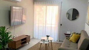 A living room with a sofa, a coffee table, a TV, a potted plant, and a window with curtains. - Apartment with an artificial beach! (Mazatlán)