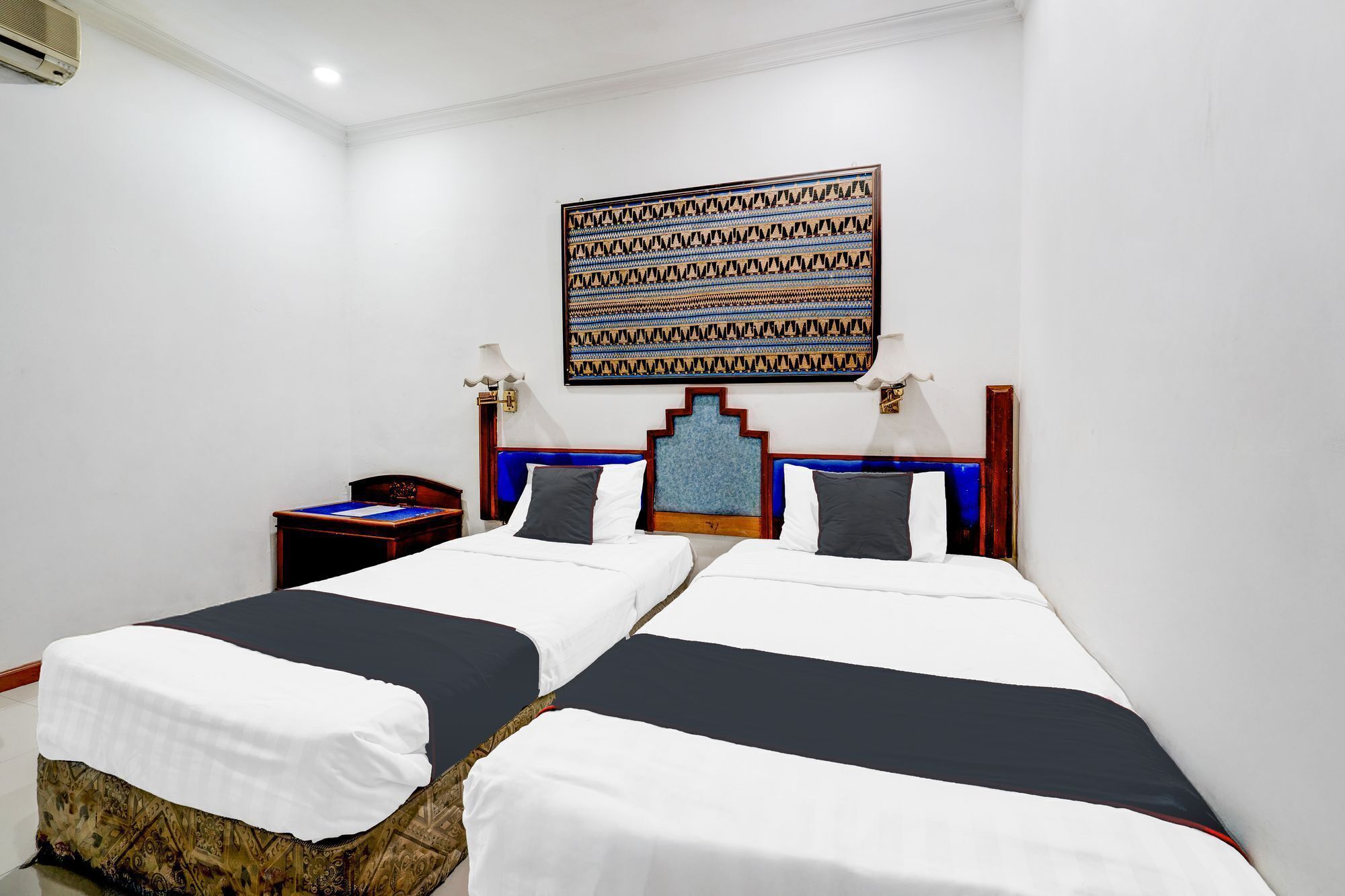 A hotel room with two beds, a patterned wall hanging, and a small table with a lamp.
