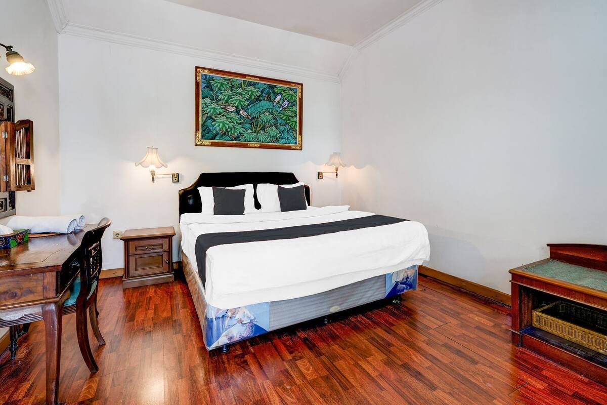 Une chambre à coucher bien rangée, avec un grand lit, deux lampes de chevet, un bureau en bois avec une chaise et un tableau accroché au mur.