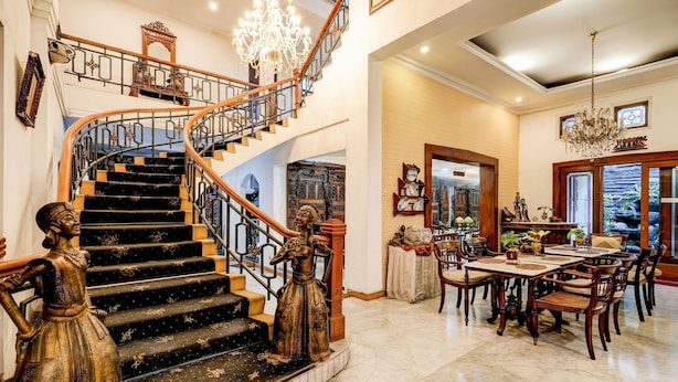 A grand staircase with ornate railings and a chandelier, leading to a dining area with wooden furniture and a view of the outdoors.