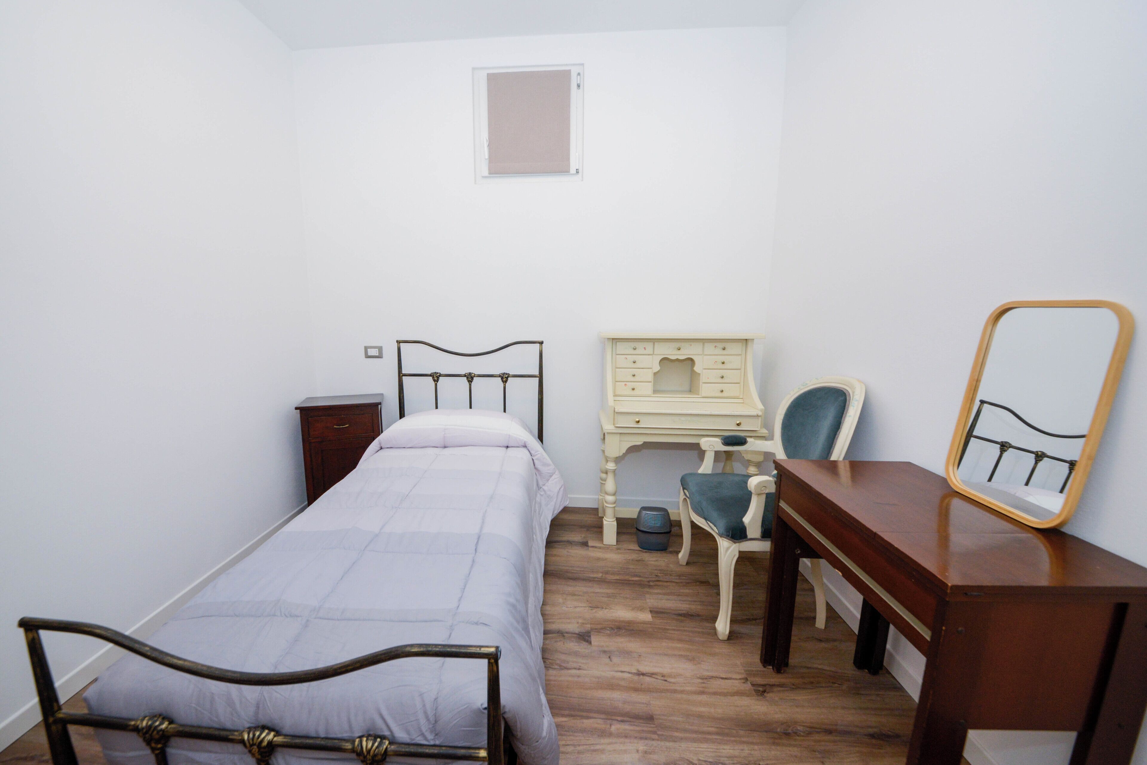 Un dormitorio con una cama individual, un aparador de madera, una silla, un escritorio pequeño y un espejo.