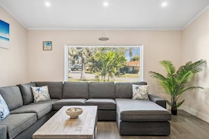 A modern living room with a sectional sofa, a coffee table, and a large window offering a view of palm trees and a building. - Florida Fun! Beach Theme, Heated Pool, Near Siesta (Sarasota)