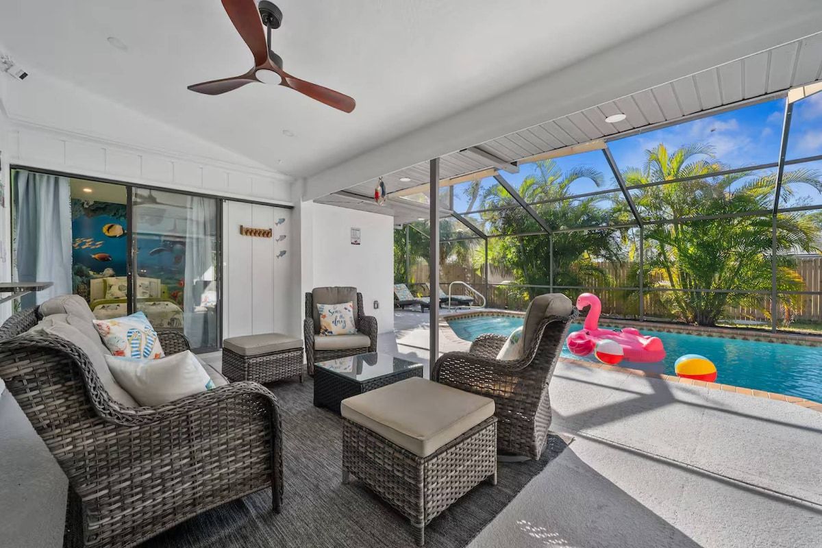 A modern indoor pool area with wicker furniture, a ceiling fan, and a view of a tropical outdoor pool.