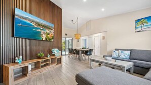 A modern living room with a large TV, wooden paneling, a dining area, and a gray sofa. - Tropical Retreat – Gulf Gate Retreat w/ Heated Pool, 7 Beds, Near Siesta Key, Families/Couples Only (Sarasota)
