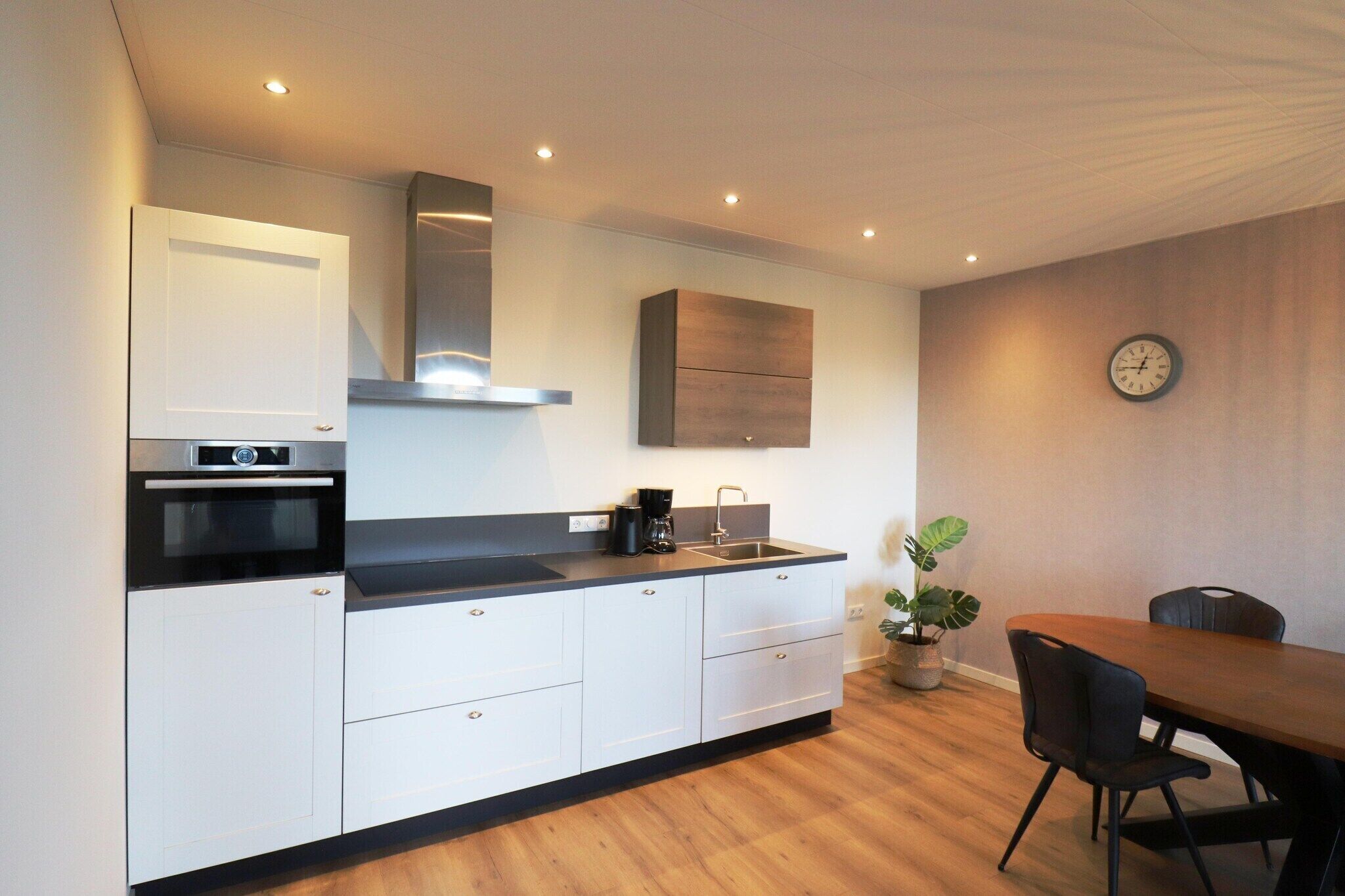 Een moderne keuken met witte kasten, een zwart aanrecht, een ingebouwde oven en een roestvrijstalen afzuigkap. Er staat een eettafel met stoelen en een potplant.