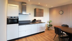 Une cuisine moderne avec des armoires blanches, un comptoir noir, un four encastré et une hotte de cuisinière en acier inoxydable. On y trouve une table à manger avec des chaises et une plante en pot.