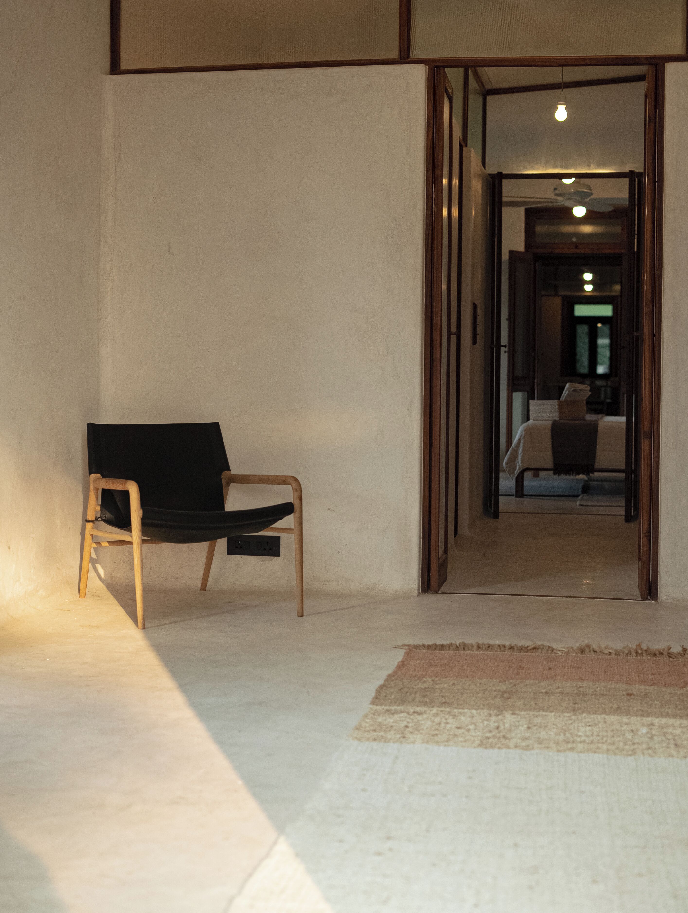 A room with a wooden chair, a small rug, and a doorway leading to another room with a bed and a ceiling fan.