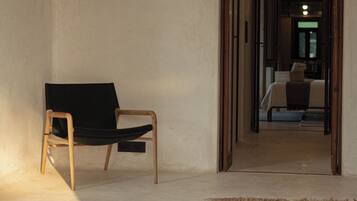 A room with a wooden chair, a small rug, and a doorway leading to another room with a bed and a ceiling fan.