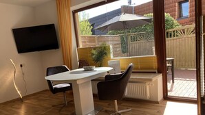 A modern living room with a dining table, two chairs, a TV mounted on the wall, and a large window with an umbrella. - The "Seebär" in Haus Meeresglück, close to the beach and central! - House Meeresglück (Dahme)