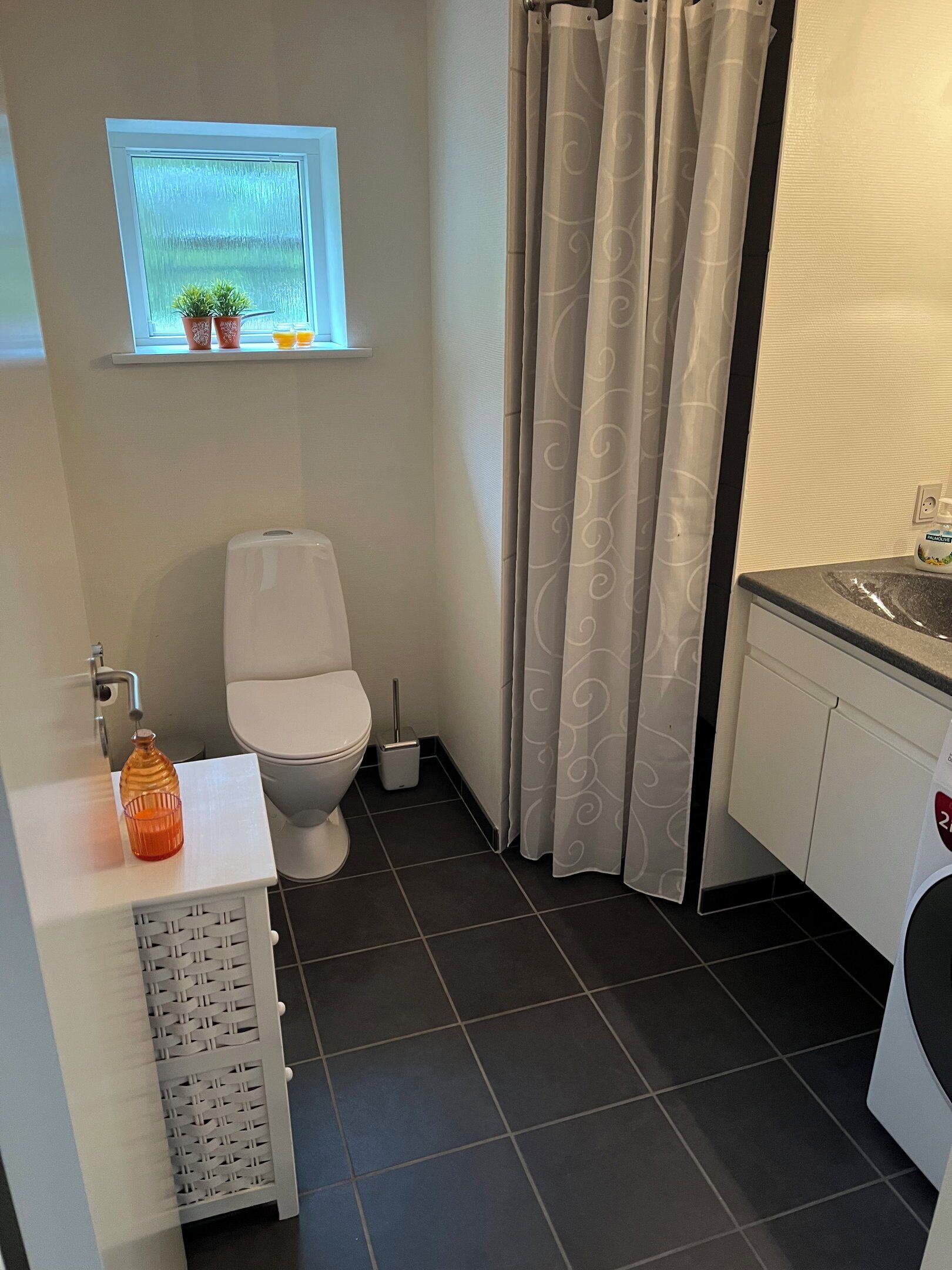 Une salle de bain avec des toilettes, une fenêtre ornée de plantes, un lavabo et un rideau de douche.