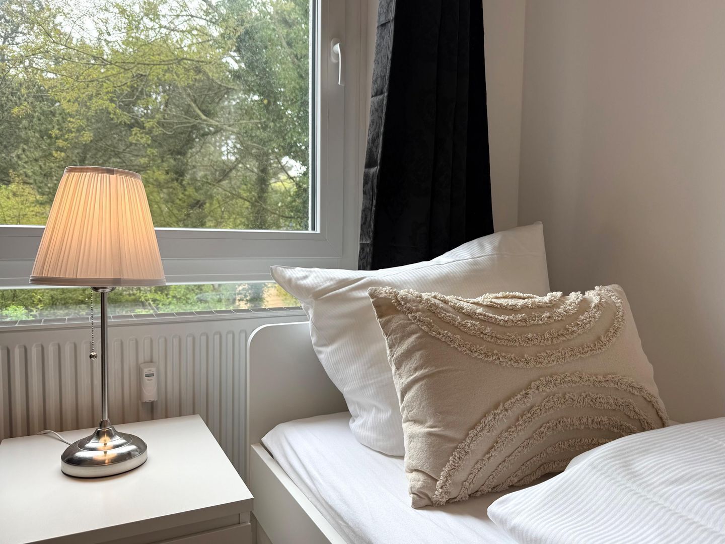 Ein Schlafzimmer mit einem Bett, einem Nachttisch mit Lampe, einem Fenster und einer Heizung.