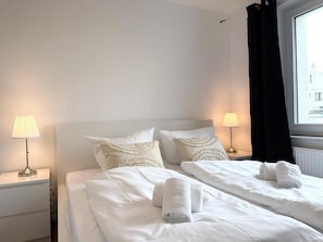 Ein Hotelzimmer mit zwei Betten, weißer Bettwäsche, dekorativen Kissen, Nachttischlampen und einem Fenster mit schwarzen Vorhängen.