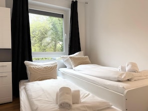Ein Schlafzimmer mit zwei Betten, einem Fenster mit Vorhängen, einem weißen Kleiderschrank und Blick auf Bäume.