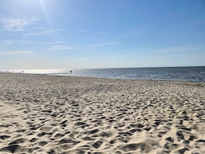 Ein sandiger Strand mit Fußspuren, die zum Wasser führen, ein klarer Himmel und ein Pier in der Ferne.