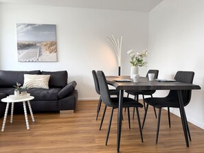 Ein modernes Wohnzimmer mit einer schwarzen Couch, einem Holzespeisesaal mit schwarzen Stühlen, einem kleinen weißen Beistelltisch und einem Bild von einer Strandansicht.