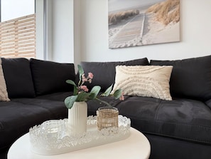 Ein modernes Wohnzimmer mit einer dunkelbraunen Ledersofa, einem weißen Beistelltisch und Dekorationselementen, darunter eine Vase mit rosa Blumen und ein gewebter Korb.