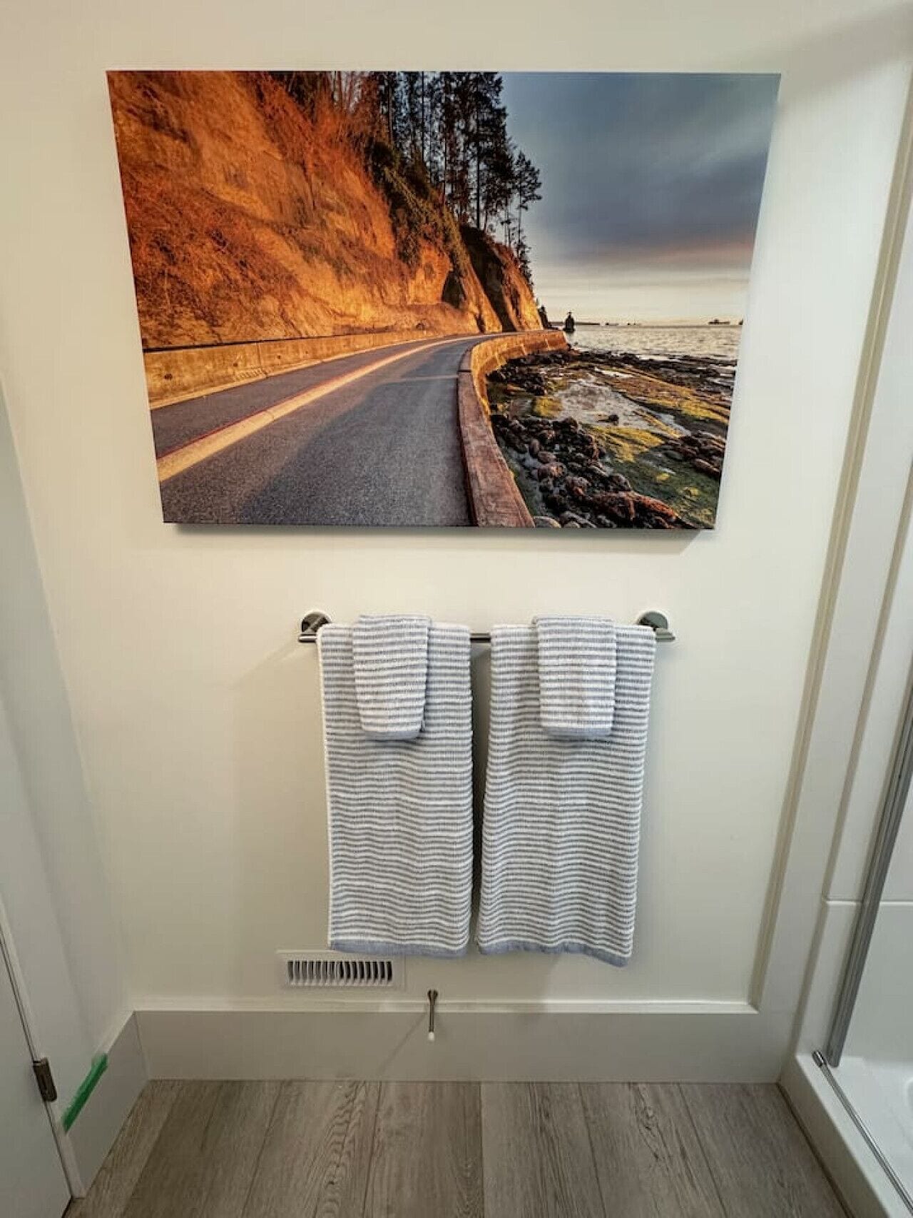 A bathroom with a painting of a coastal road and two striped towels hanging on a rack.