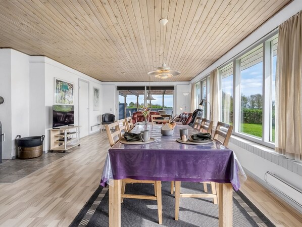 Enjoy meals in this dining area with a long table, high ceiling, and large windows offering a view of the outdoors.