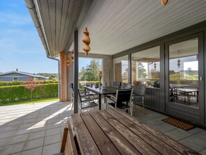 En täckt uteplats med ett träbord och stolar, en bänk och en glasdörr som leder ut till ett utomhusområde.