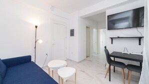 Un salon moderne avec un canapé bleu, deux petites tables d’appoint blanches, un coin repas avec une table et des chaises noires, et un téléviseur fixé au mur.