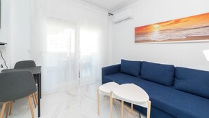 Un salon moderne avec un canapé bleu, une table blanche et un fauteuil. On y trouve une grande fenêtre avec des rideaux légers, un climatiseur et un tableau représentant un coucher de soleil sur l’océan.