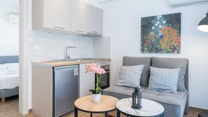 En kompakt opholdsstue med et tekøkken, en sofa med puder, et lille rundt bord og en potteplante.