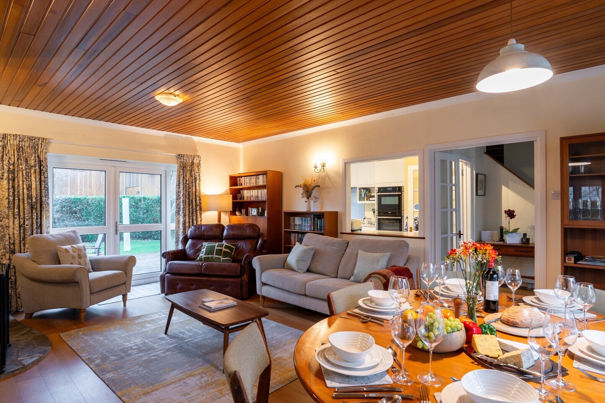 A cozy living room with a wooden ceiling, a fireplace, a dining area set for a meal, and a bookshelf.